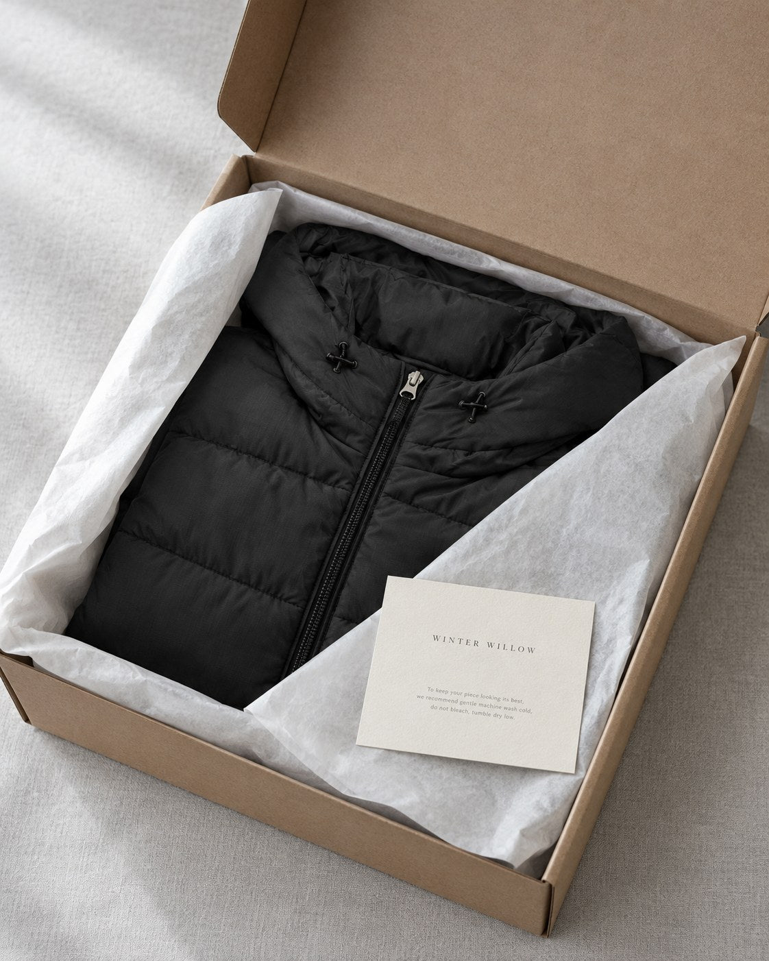 A coat folded in recycled tissue inside a plain kraft box, with a care card